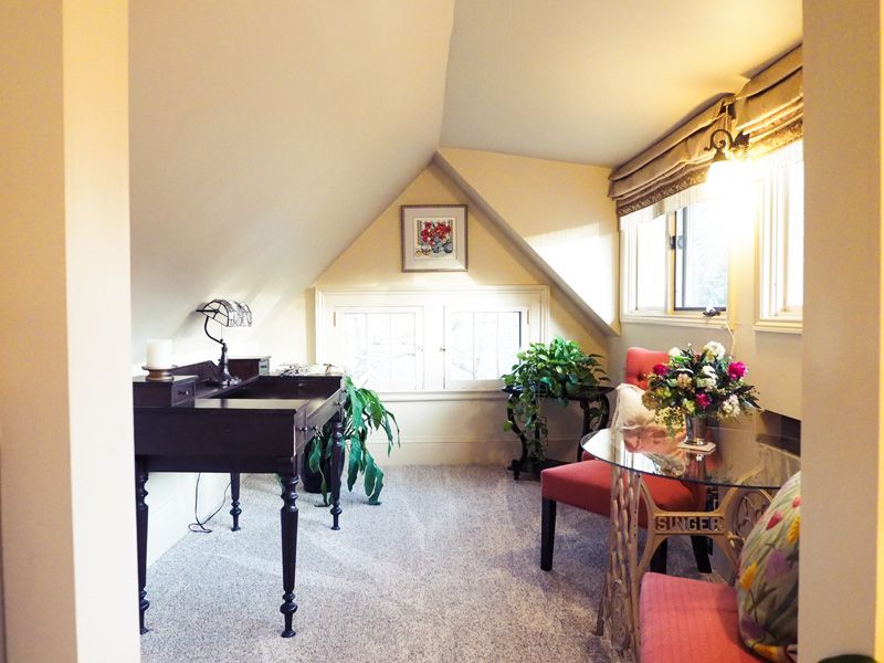 A sitting room in the attic with a desk, two red armchairs, a standing lamp and several plants with windows on the whole right wall.