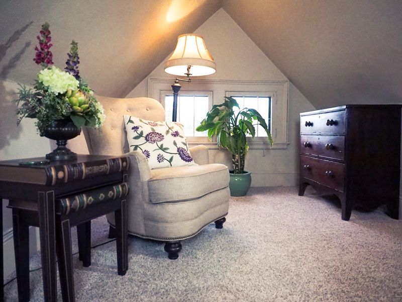 A relaxing area at one of the suites in the Asheville bed and breakfast. An armchair with a dark wooden dresser, a bright carpet, a little side table and a flower bouquet.