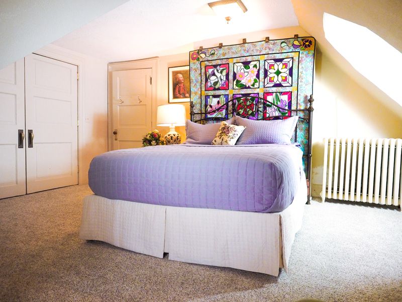 A bright, beige room with a queen bed with purple linen and a quilt hanging on the wall at the head of the bed.