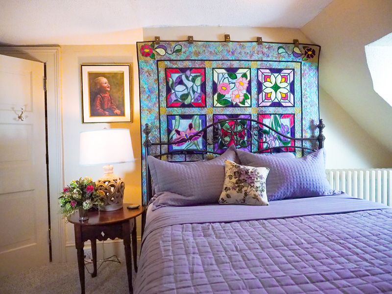 A bright, beige room with a queen bed with purple linen and a quilt hanging on the wall at the head of the bed.