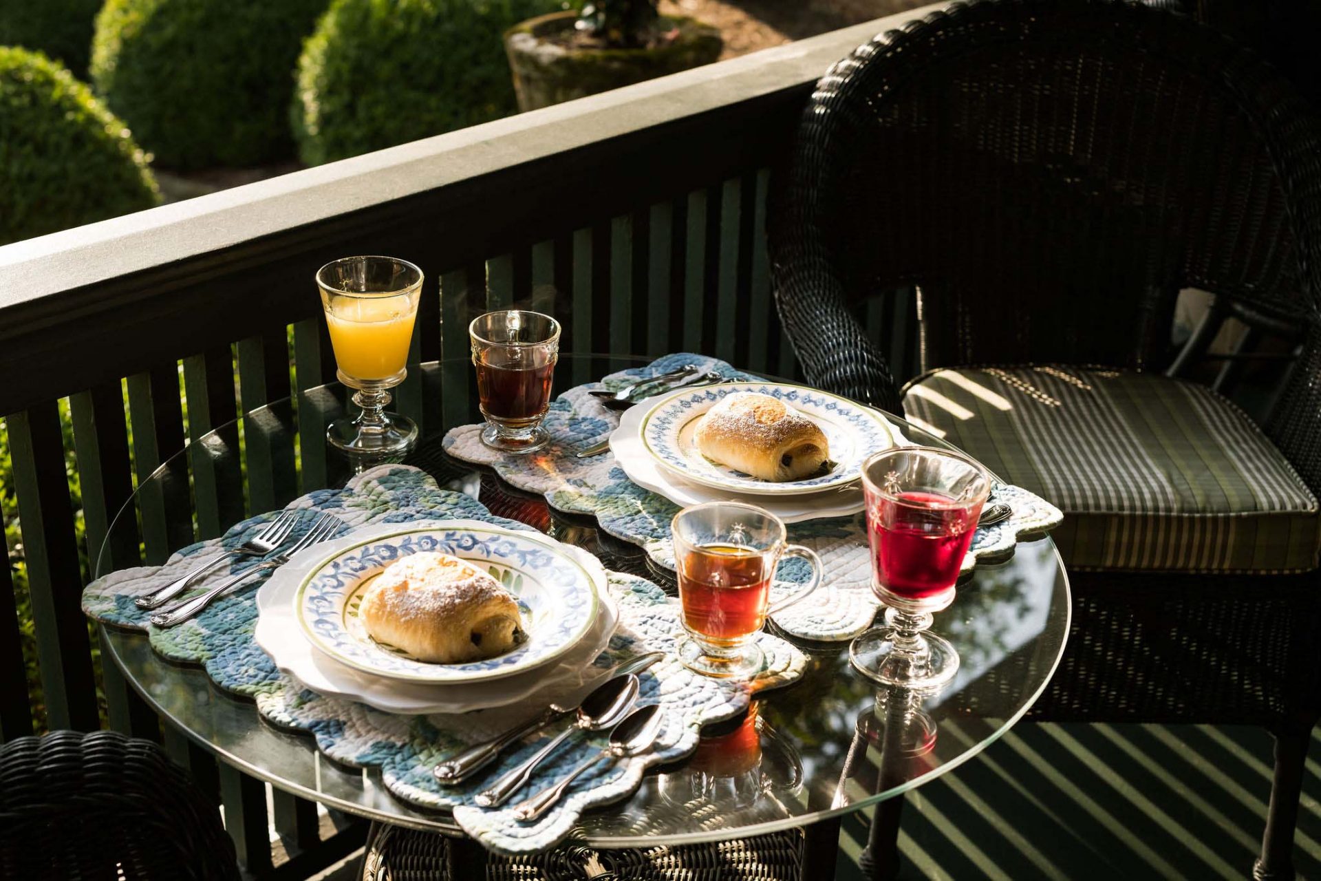 Breakfast served on the porch. A table for two with floral placemats with ceramic dishes and cuttlery, two glasses of juice, two glasses of tea and two home-baked buns.