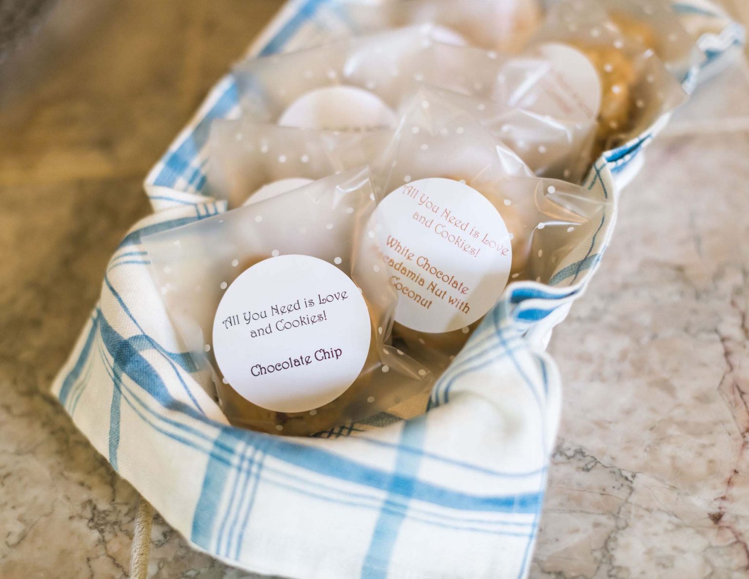 A close-up of chocolate chip cookies individually packed in foil, in a woven basket with a white-blue cloth.