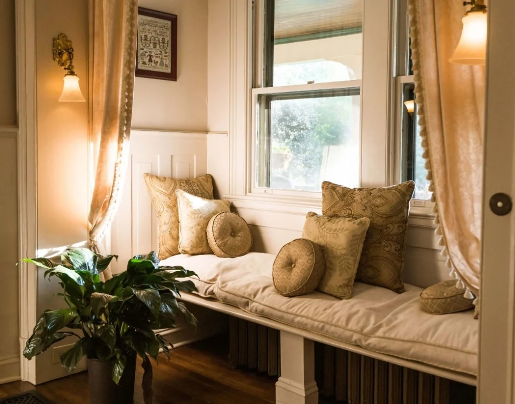 A nook with a cushioned bench and decorative pillows, beneath a window with draped curtains.