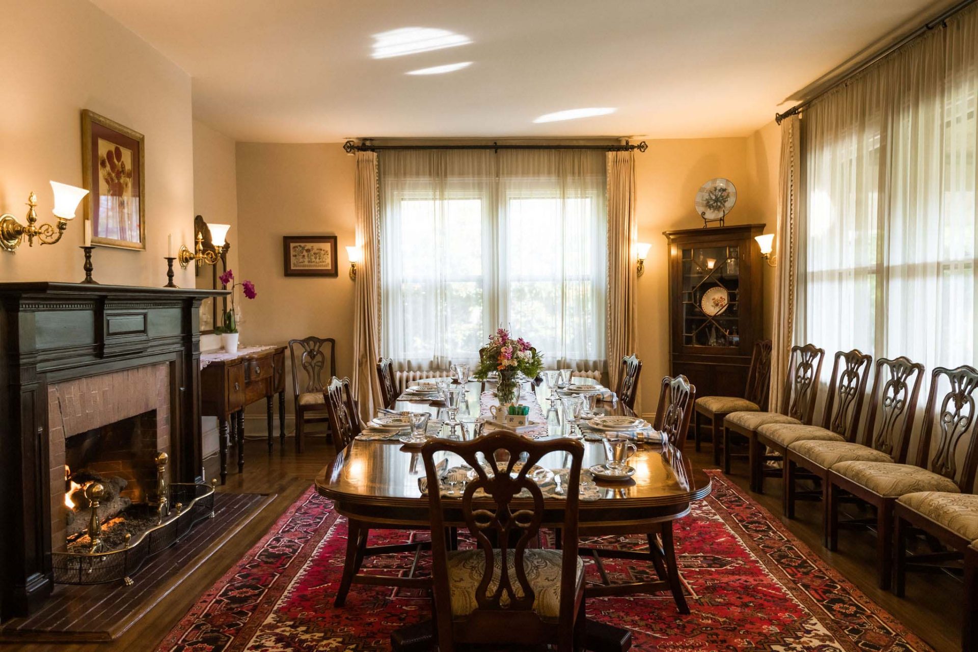 A spacious dining room area with a big, dark wood dining table with wooden chairs, a fireplace on the sidewall, a dark wood dresser with decorative ceramics in the corner. Big windows with sheer curtains.