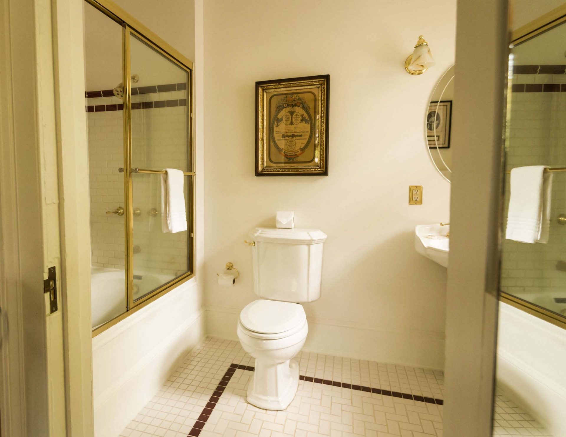 A beige-gold bathroom with a glass-door bathtub; a toilet, and a sink with an oval mirror. A gold painting hanging on the wall.
