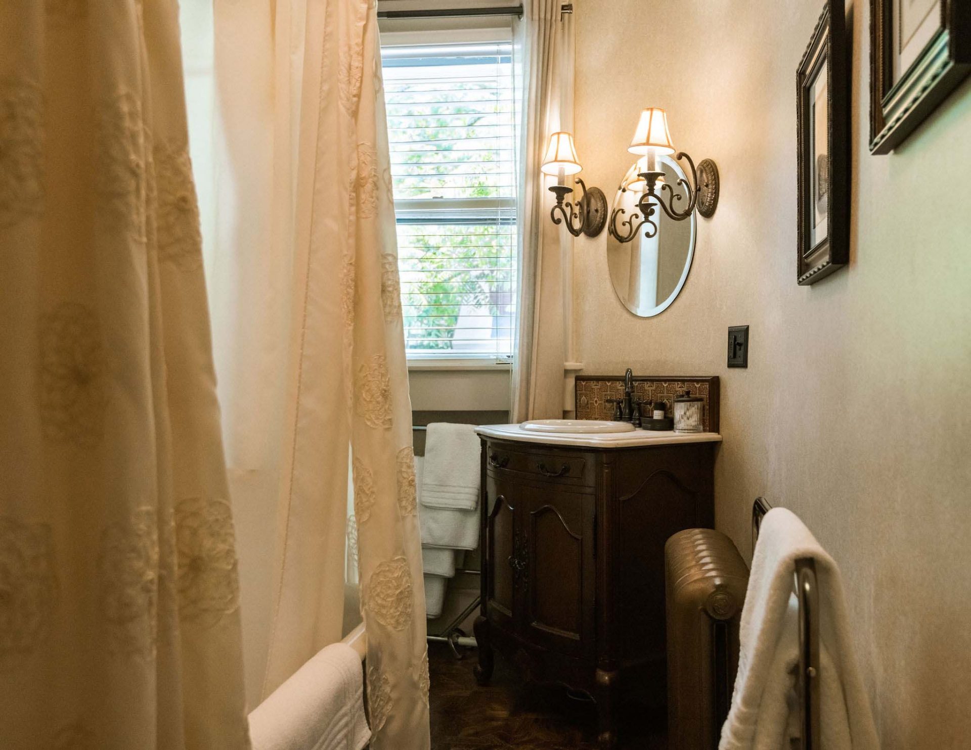 An oval shaped, vintage bathroom with a claw foot tub with shower above and a creamy curtain, sink in a dark wood dresser, and a mirror.