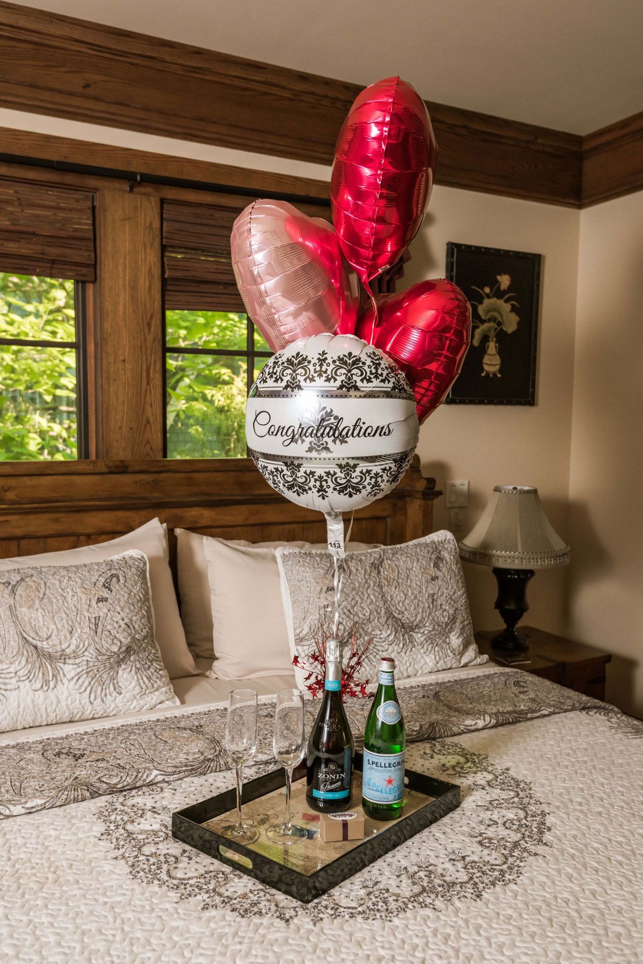 White and red heart-shaped balooons tied to a tray with a prosecco bottle, water bottle, chocolate truffles, and champagne glasses, arranged on a king bed.