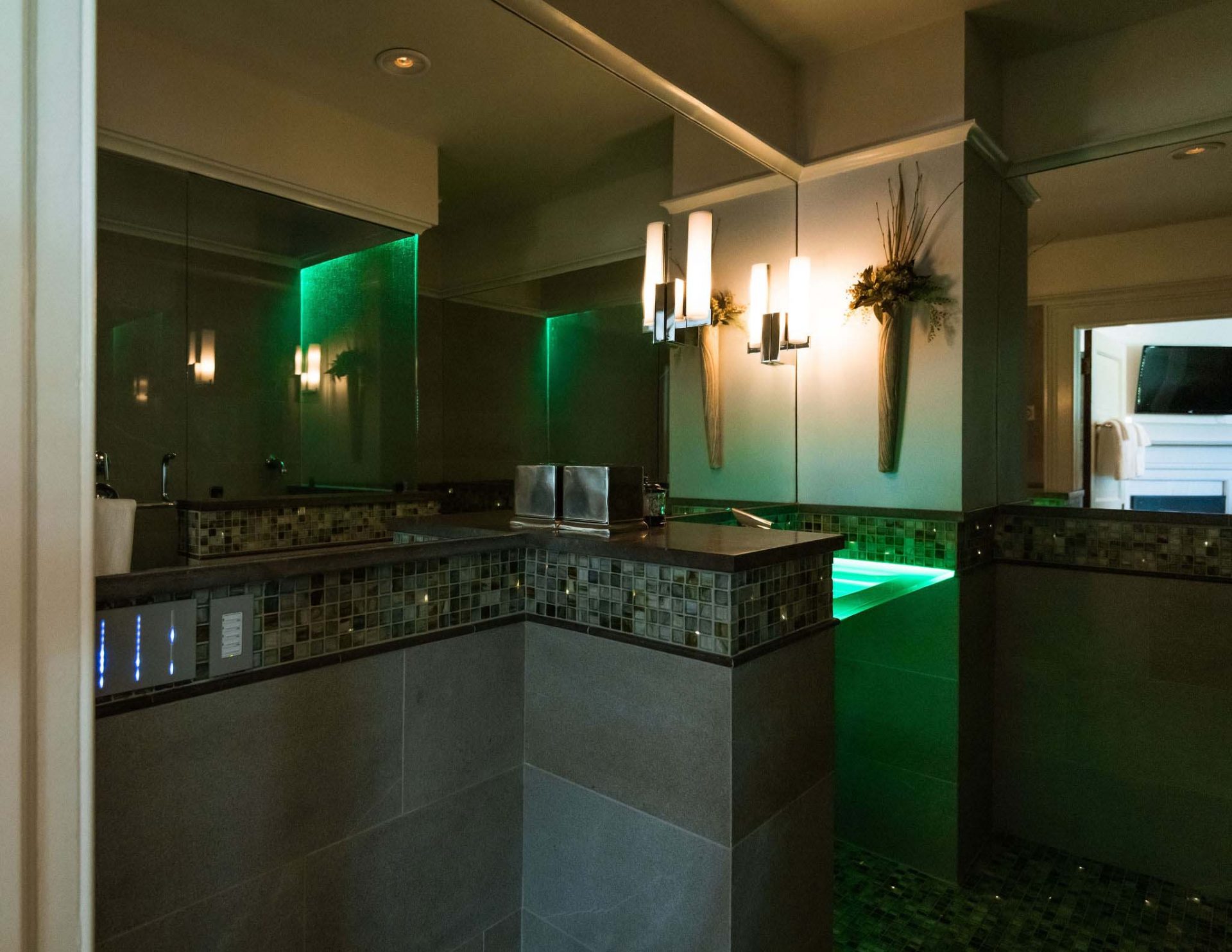 A luxurious bath with colorful fiber optic lighting and a green-lit glass sink, with dimmed lights. Long mirror on the whole wall above the sink.
