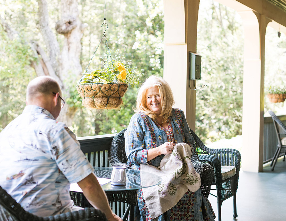 The Innkeepers sitting together the porch. Shawnie is weraing a long, floral, blue dress, and laughs while embroidering.