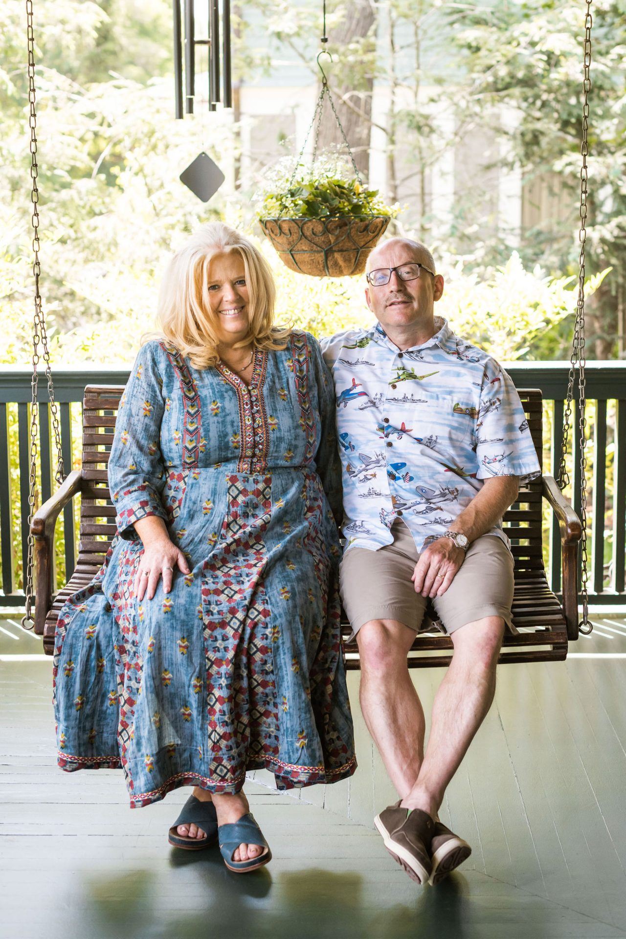 The Innkeepers sitting together on a swing on the porch and smiling. Shawnie weraing a long, floral dress, Willy wearing khaki shorts and a colorful, airplane-patterned shirt.