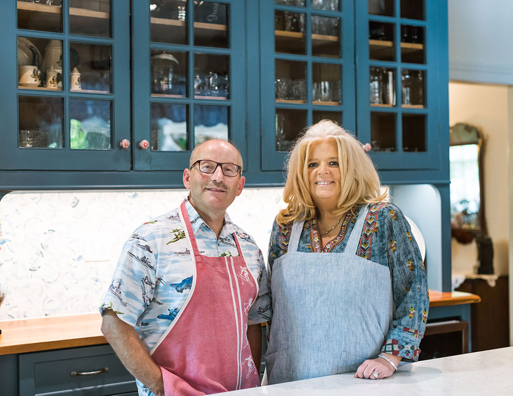 The Innkeepers standing together in the kitchen at a table. Both wearing aprons and smiling, blue cabinets with glass fronts in the background.