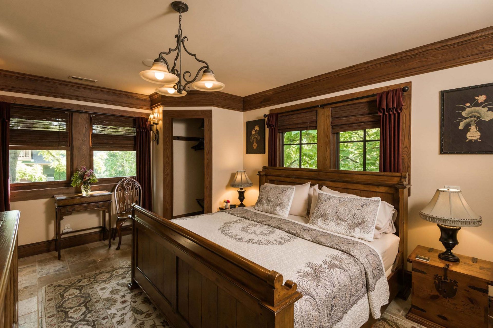 A dark wooden king bedroom with bing windows on two walls, a walk-in closet, two night stands and lamps.