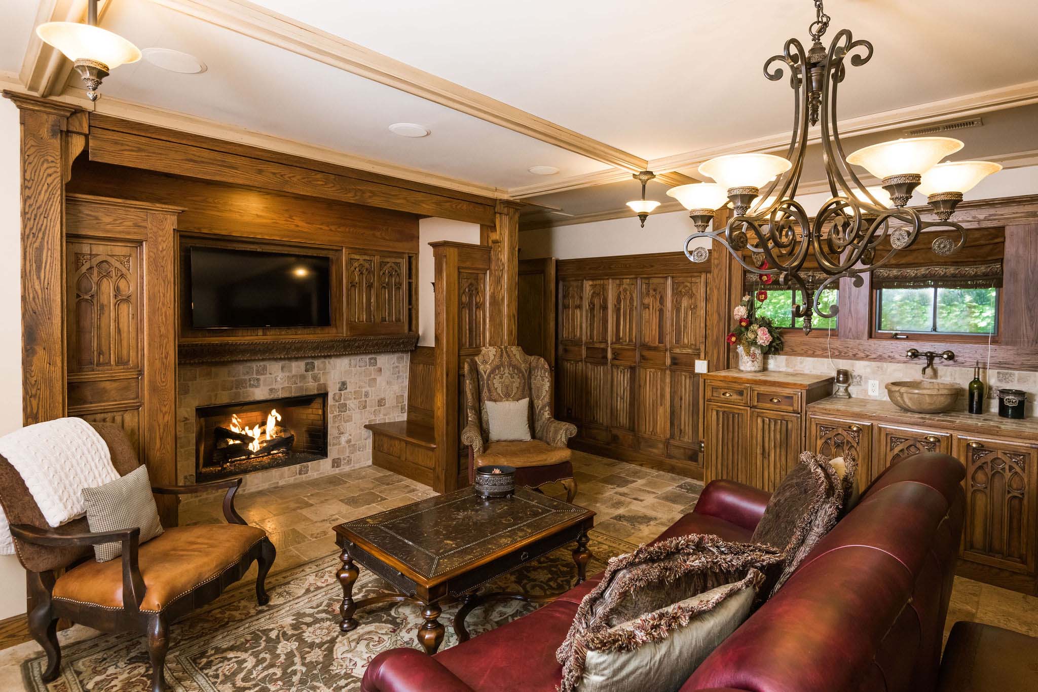 A sitting room with hand-carved paneling imported from India, a fireplace, couch, two club chairs, a table in the middle, and a TV on the wall. A set of dressers at the window with a sink, below the window.