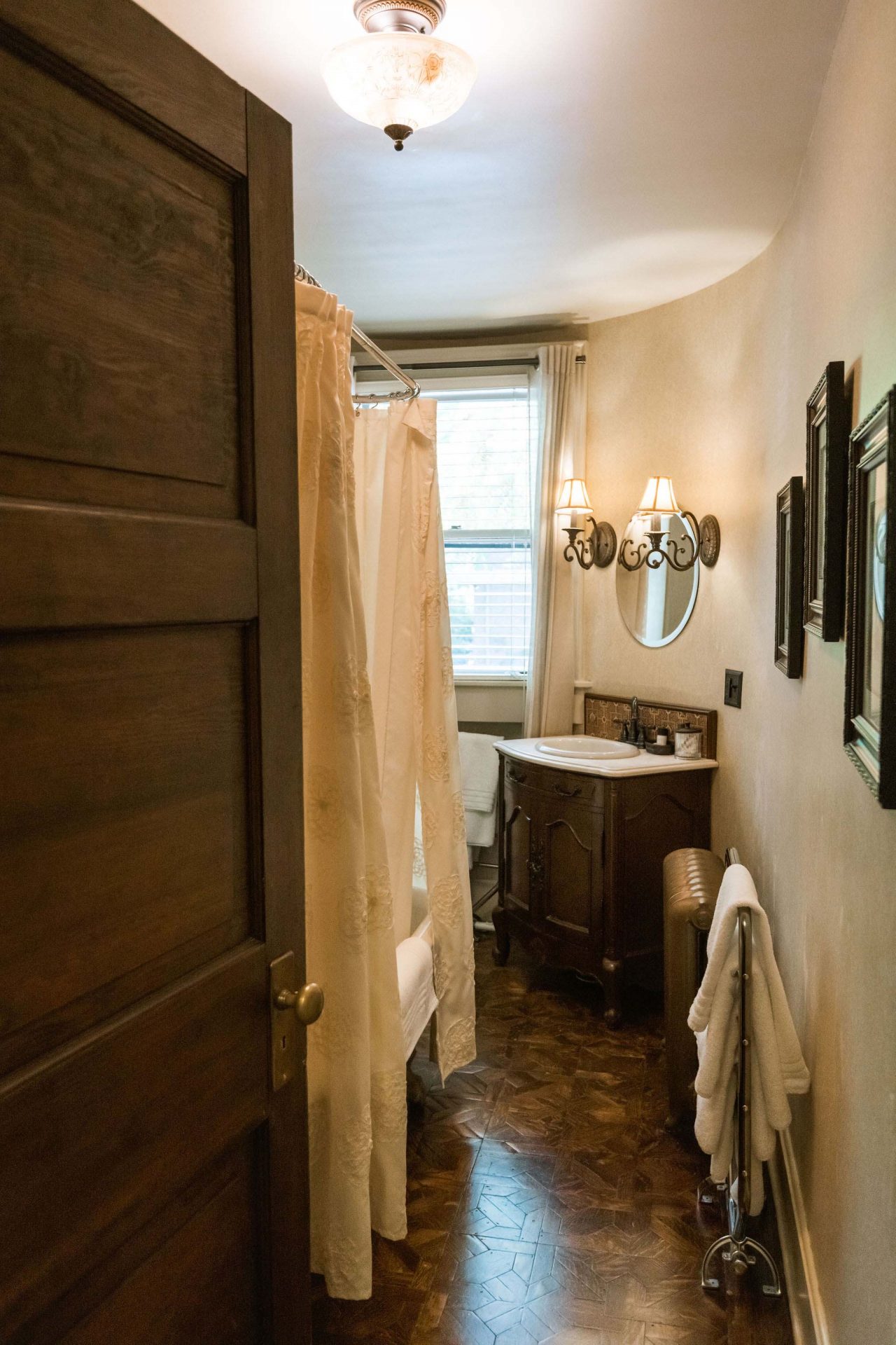 A bathroom with a claw foot tub with shower above and a creamy curtain, sink in a dark wood dresser, a mirror, and several paintings hanging over the walls.