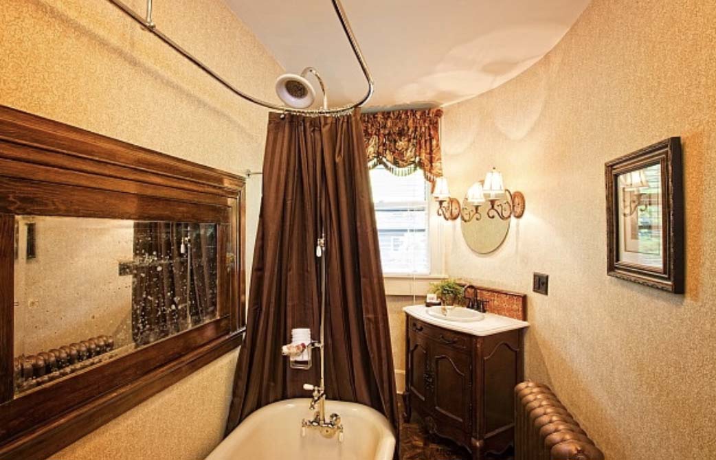 An oval shaped, vintage bathroom with a claw foot tub with shower above and a dark brown curtain, sink in a dark wood dresser, and a mirror.