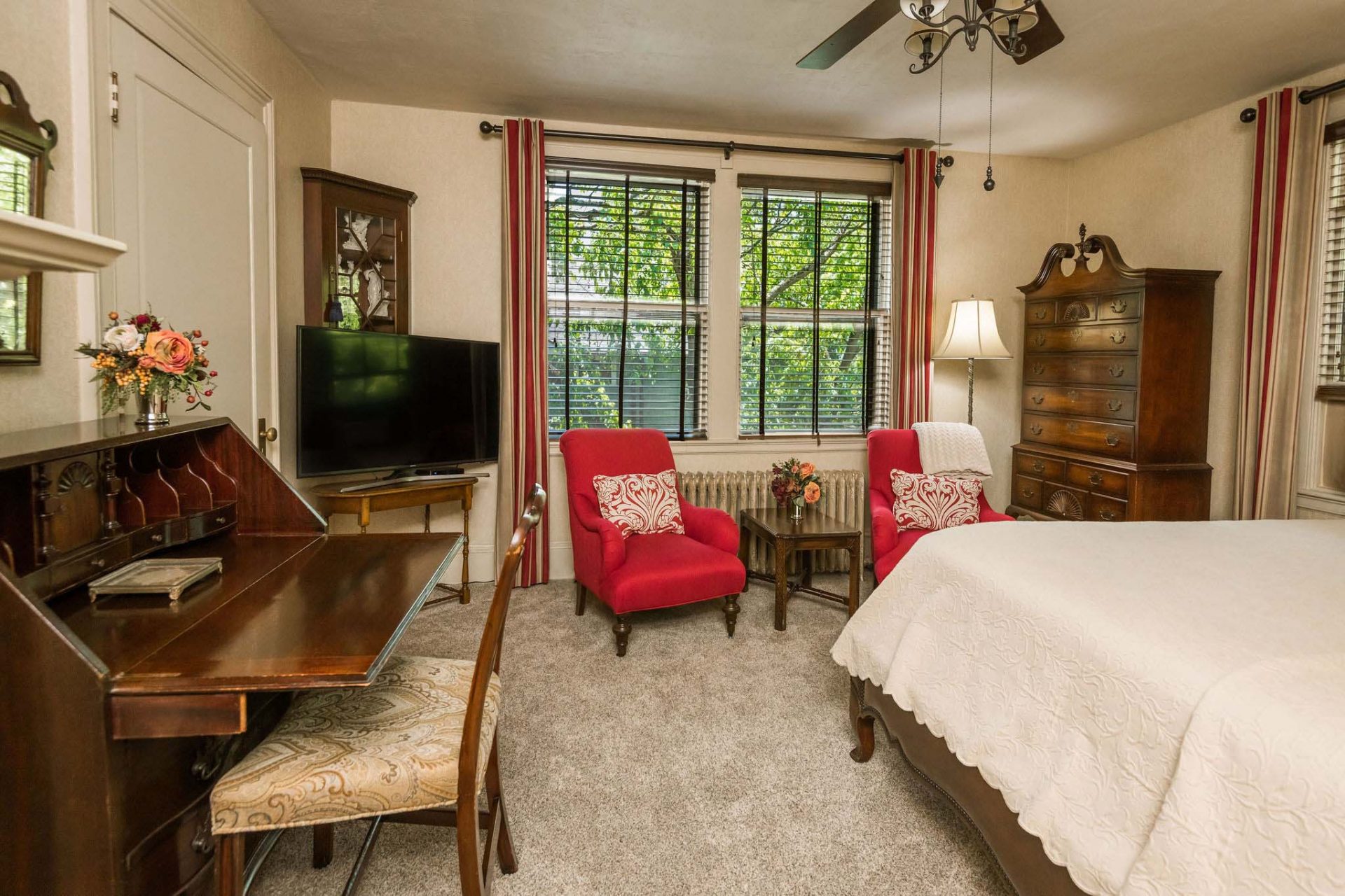A carpeted king bedroom with two red club chairs, a dark wood dresser in the corner, a TV set, and a vintage desk