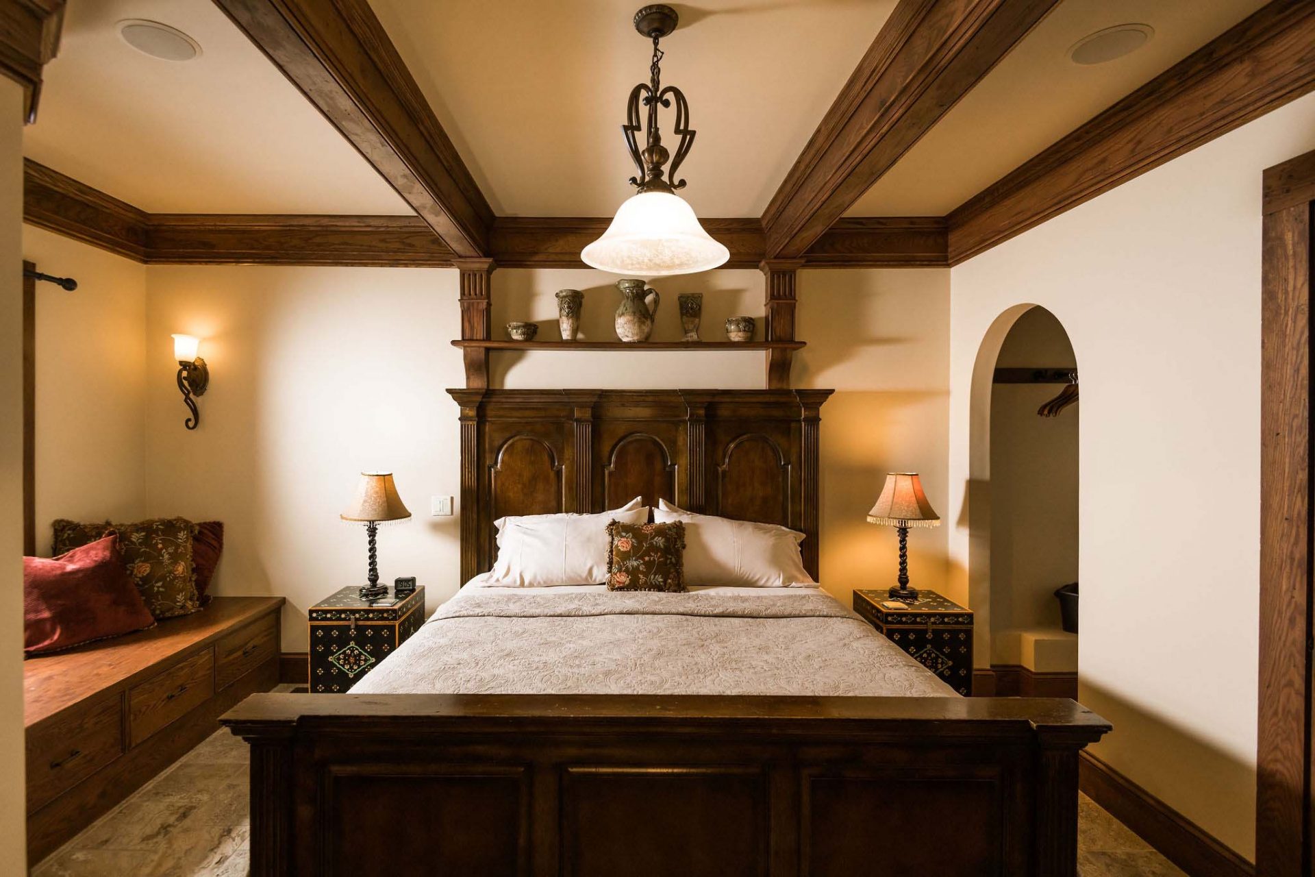 A king bedroom in dark wood with tiled floors. Broad wooden window sill with cushions, dark wooden bed frame with a decorative headboard, two ornamented night stands with lamps.