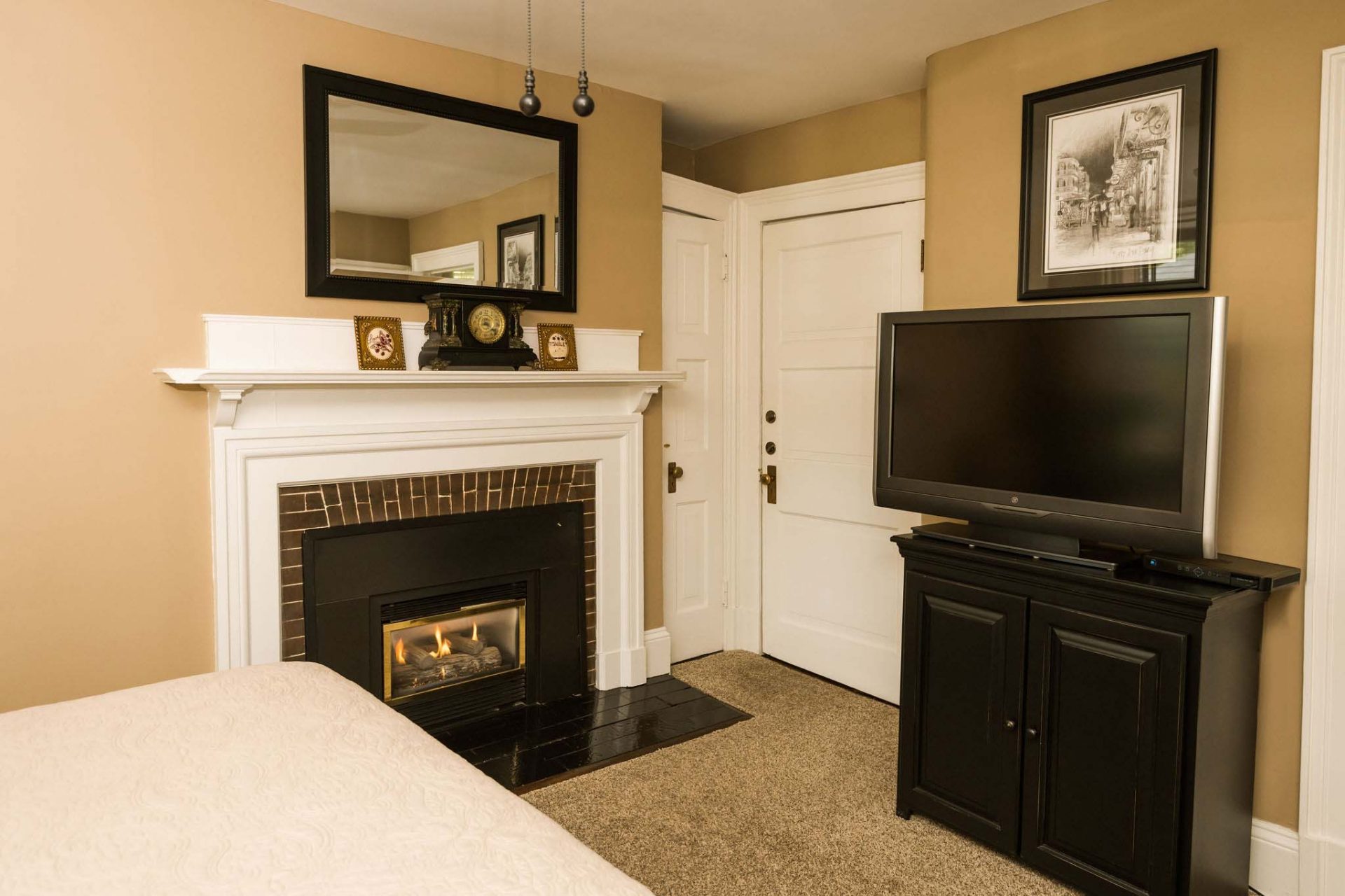 A carpeted king bedroom with a fireplace, a mirror, and a TV. Decorated with a vintage clock and several paintings.