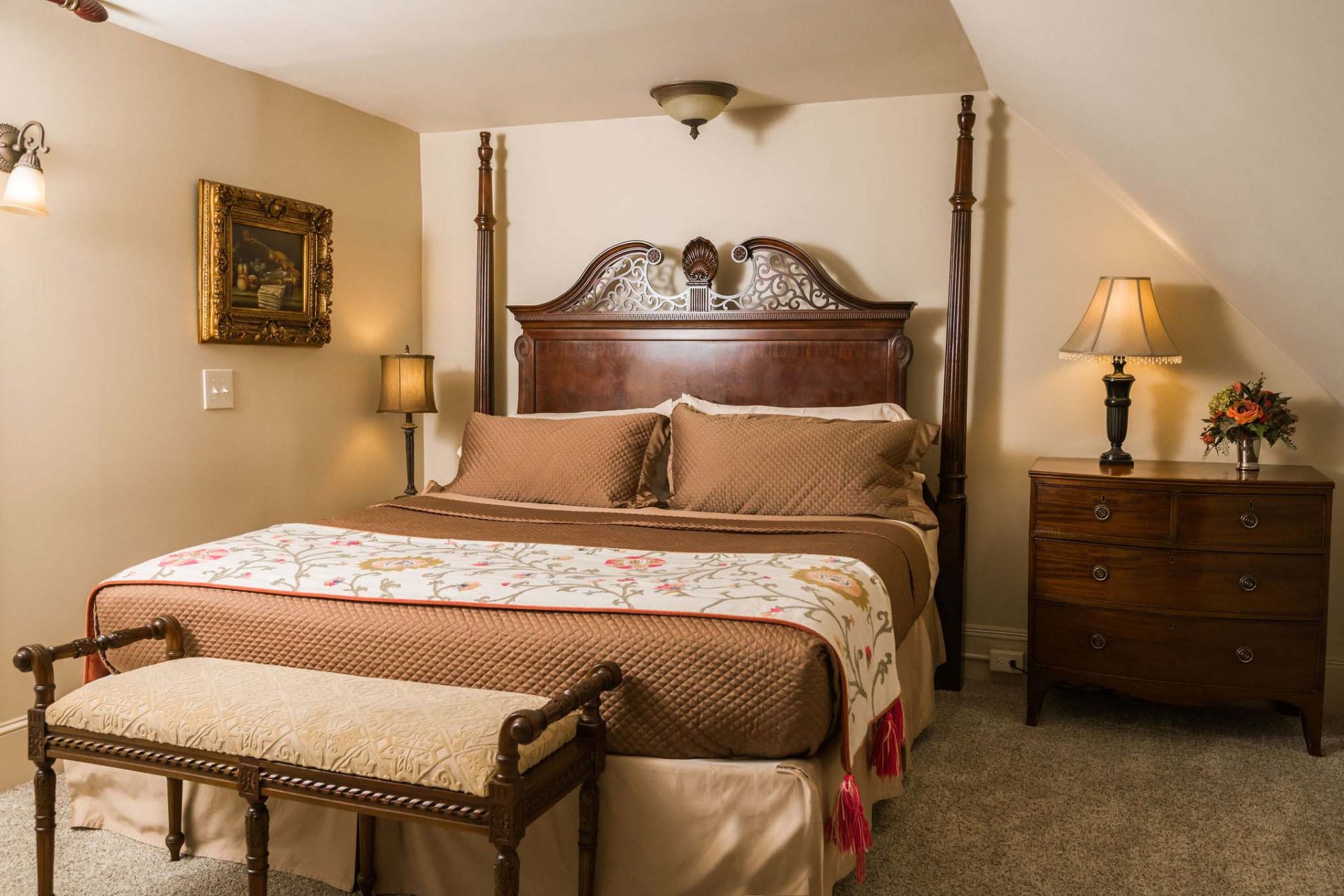 A king bedroom in a 5-room bed and breakfast suite. Kind bed with a dark wood, decorative headboard. A night stand, a cushioned bench and a night lamp. A gold-framed painting on the wall.