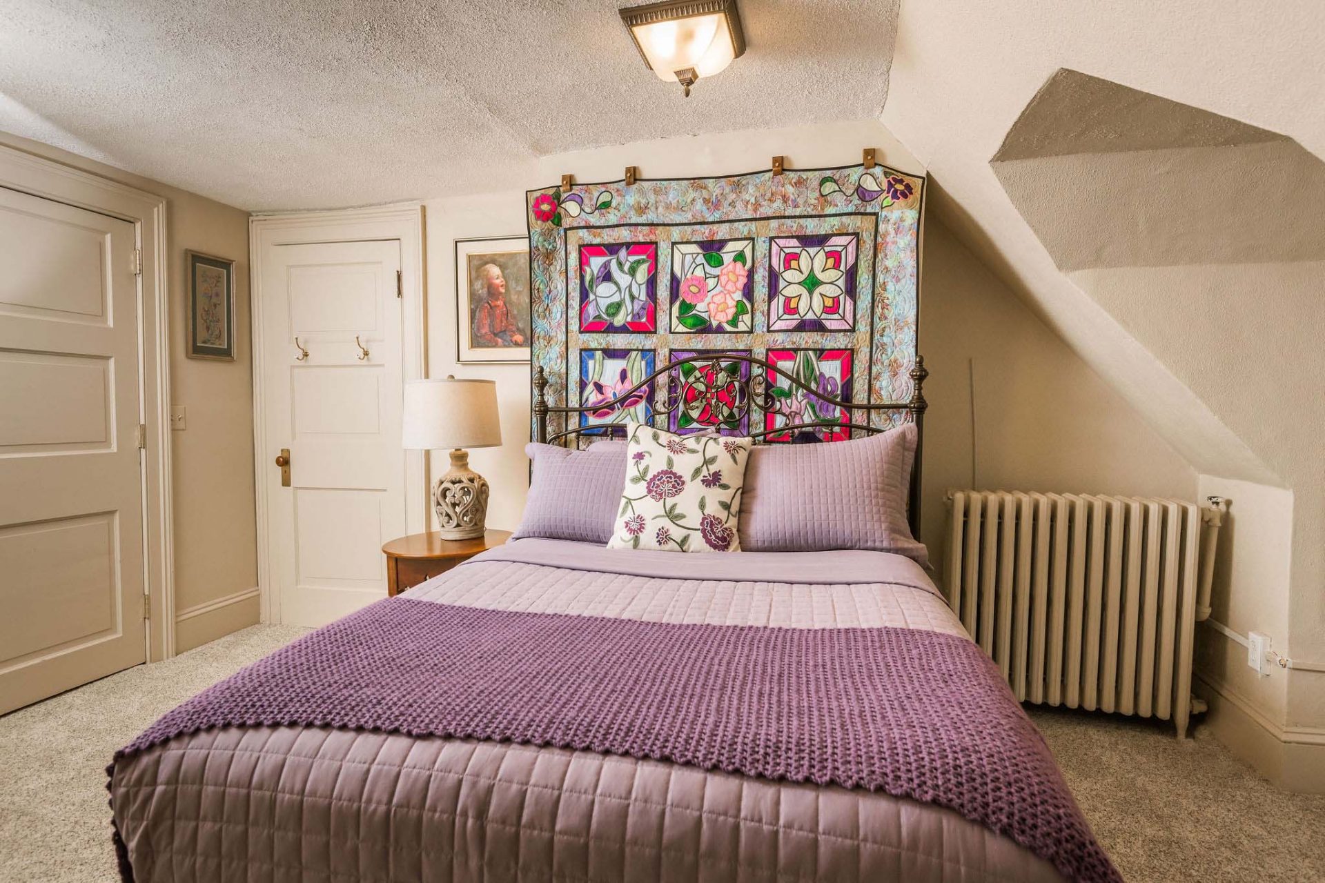 A bright, beige room with a queen bed with purple linen and a quilt hanging on the wall at the head of the bed.