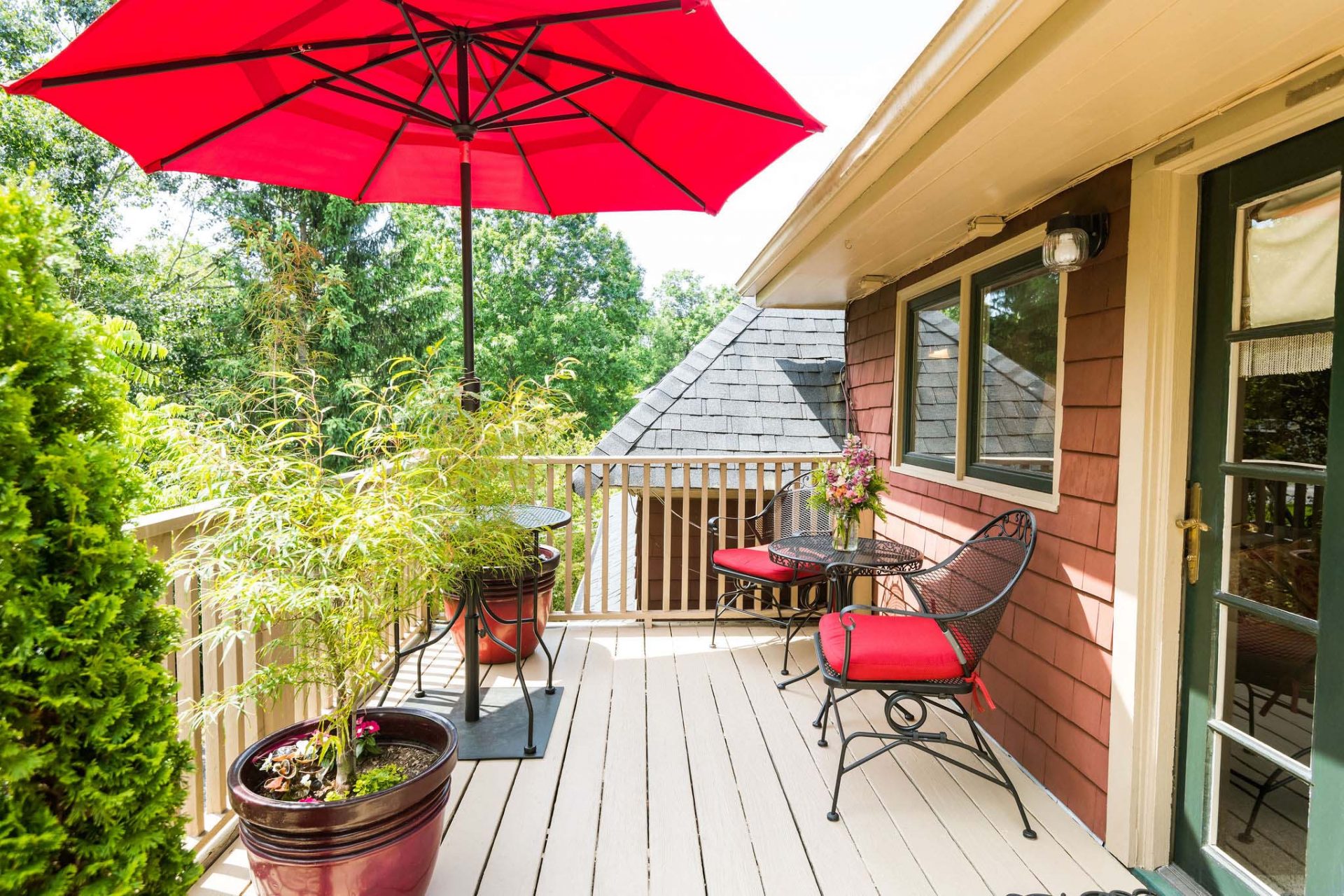 A private fenced upper deck with several plants, two chairs, small table, and a red sun garden umbrella.