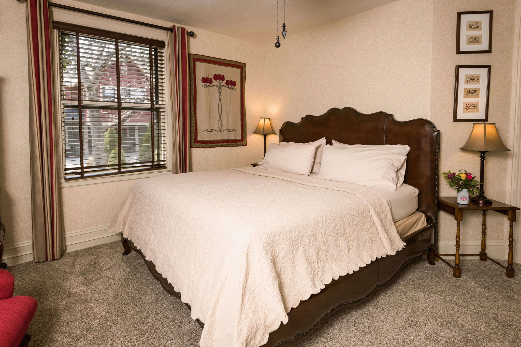 A carpeted king bedroom with two red club chairs, a dark wood dresser in the corner, two night stands with lamps and several paintings on the walls.