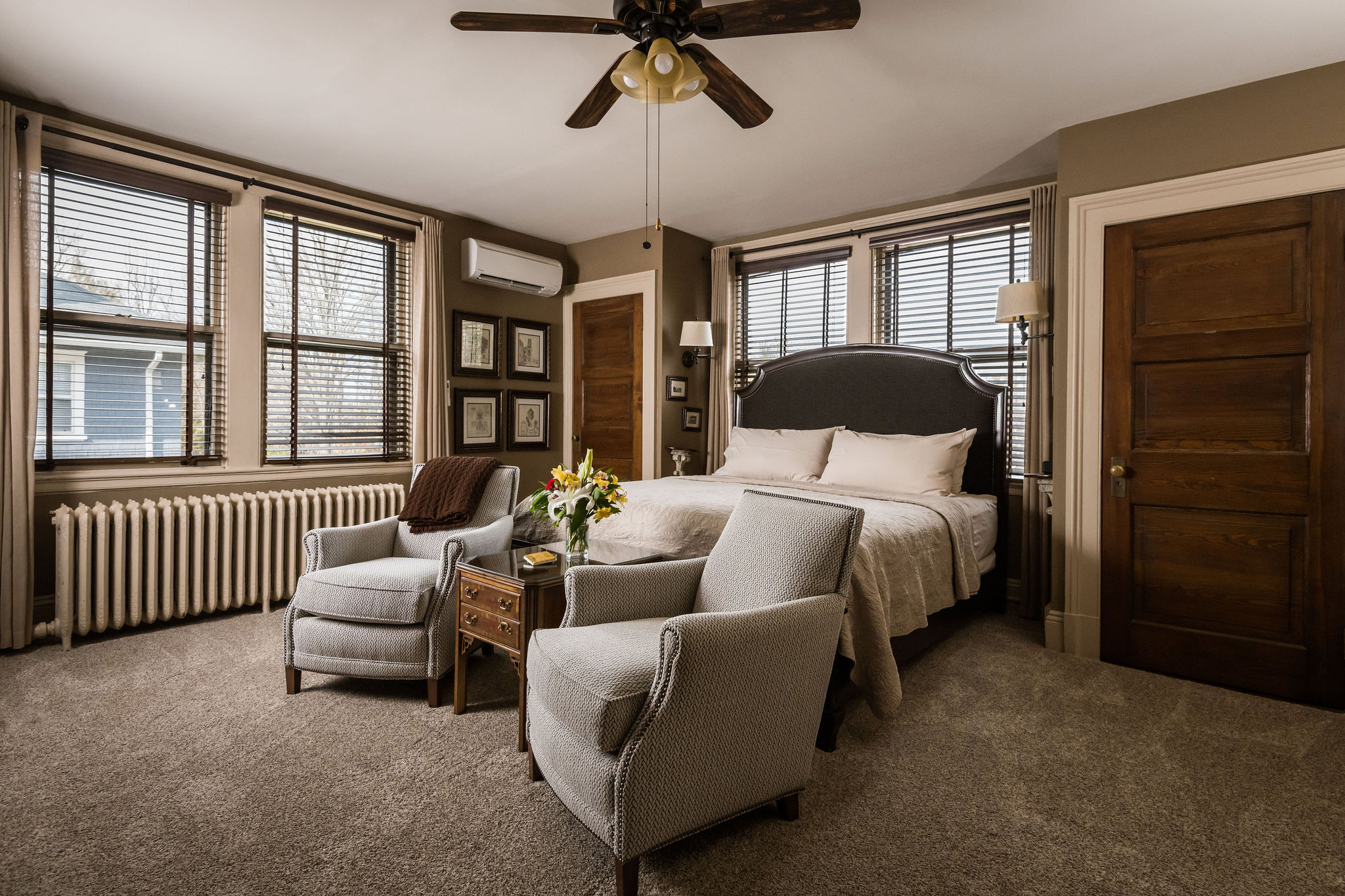 A king bedroom with a glass table, a decorative club chair and 3 dark wood doors. Two dark wood dressers with a marble countertop with 2 woven baskets below it. A gold sink on the countertop, and a long, dark-framed mirror above it.