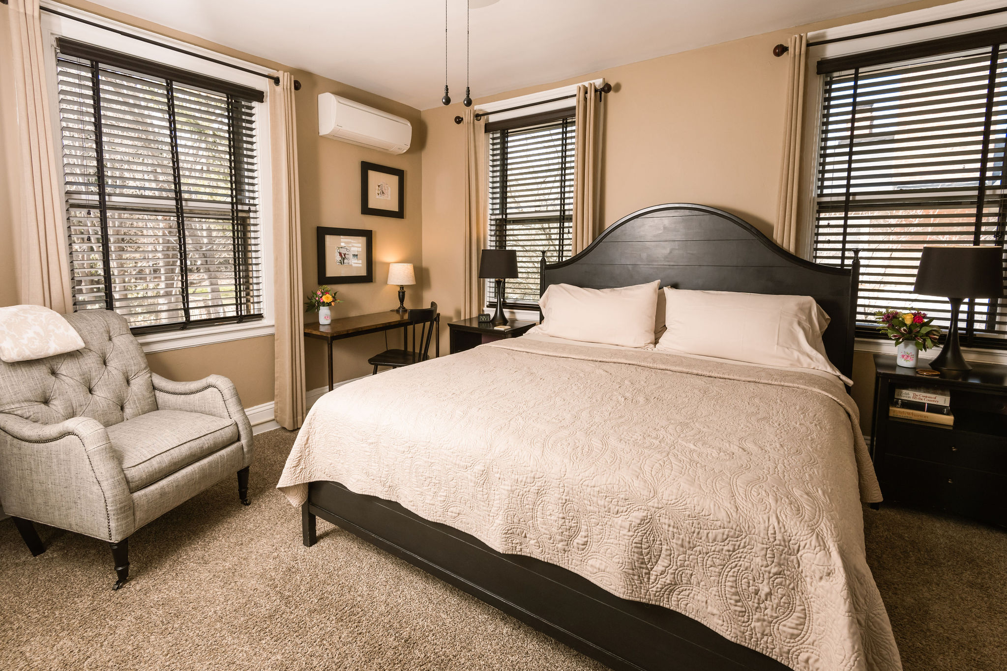 A carpeted king bedroom with beige bedding, two night stands with lamps, armchair, fan, and several paintings on the walls.